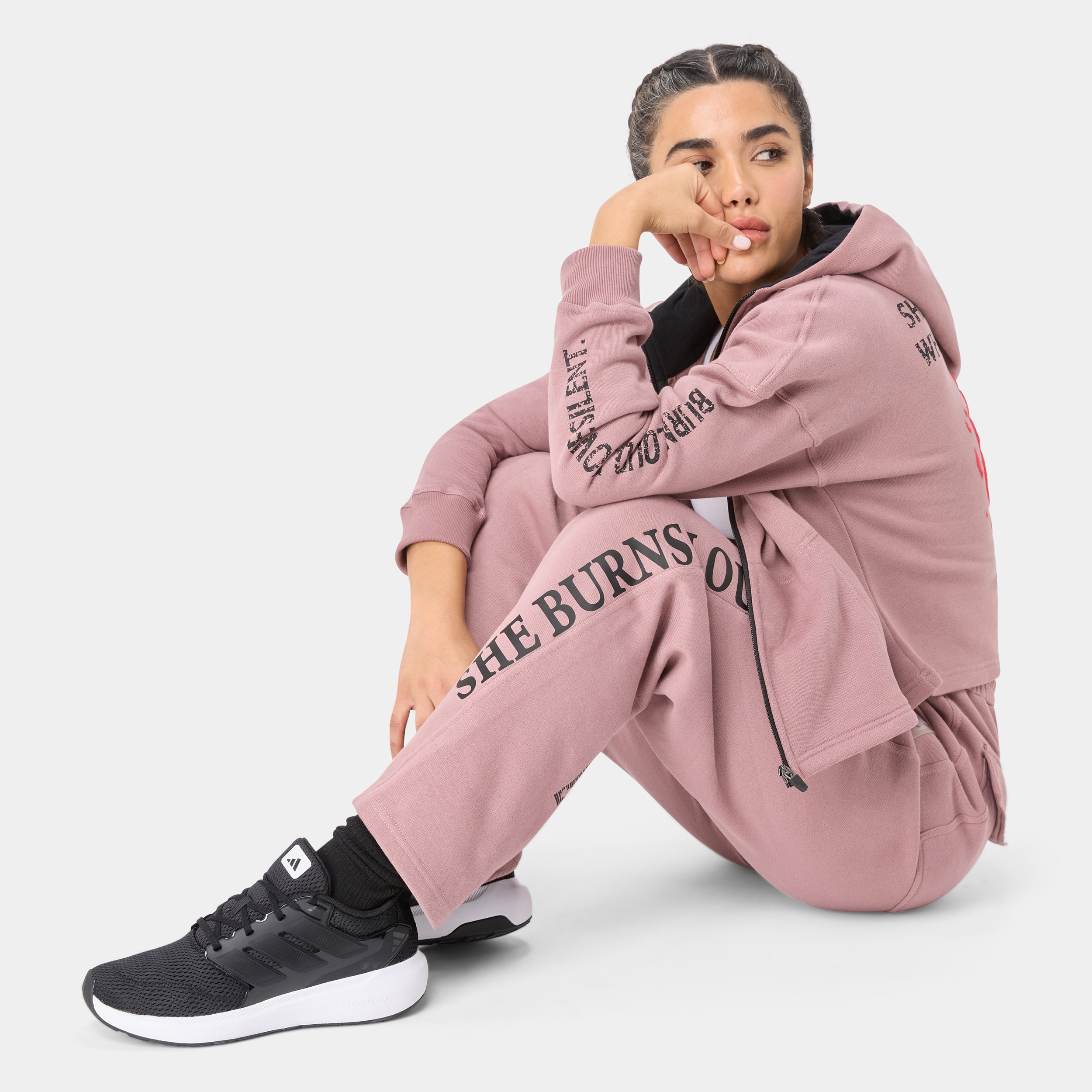 ALPHAFIRE BOTTOM — STORM ROSE — Seated floor pose close-up, pink tracksuit with SHE BURNS LOUDER leg and sleeve text