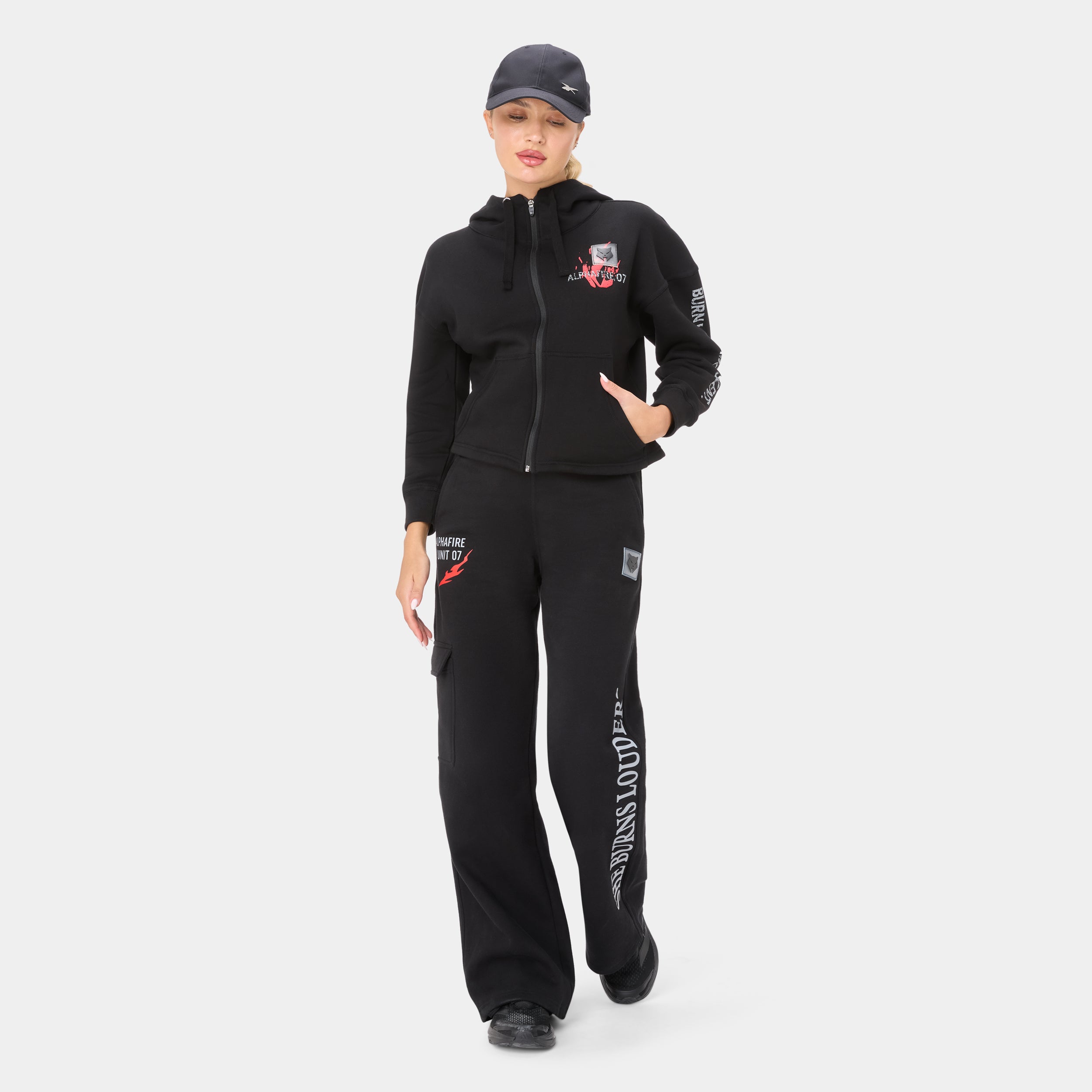 ALPHAFIRE CROP HOODIE — WAR BLACK — Full-body front ¾ walking pose, black tracksuit with cap, hand in pocket, leg text
