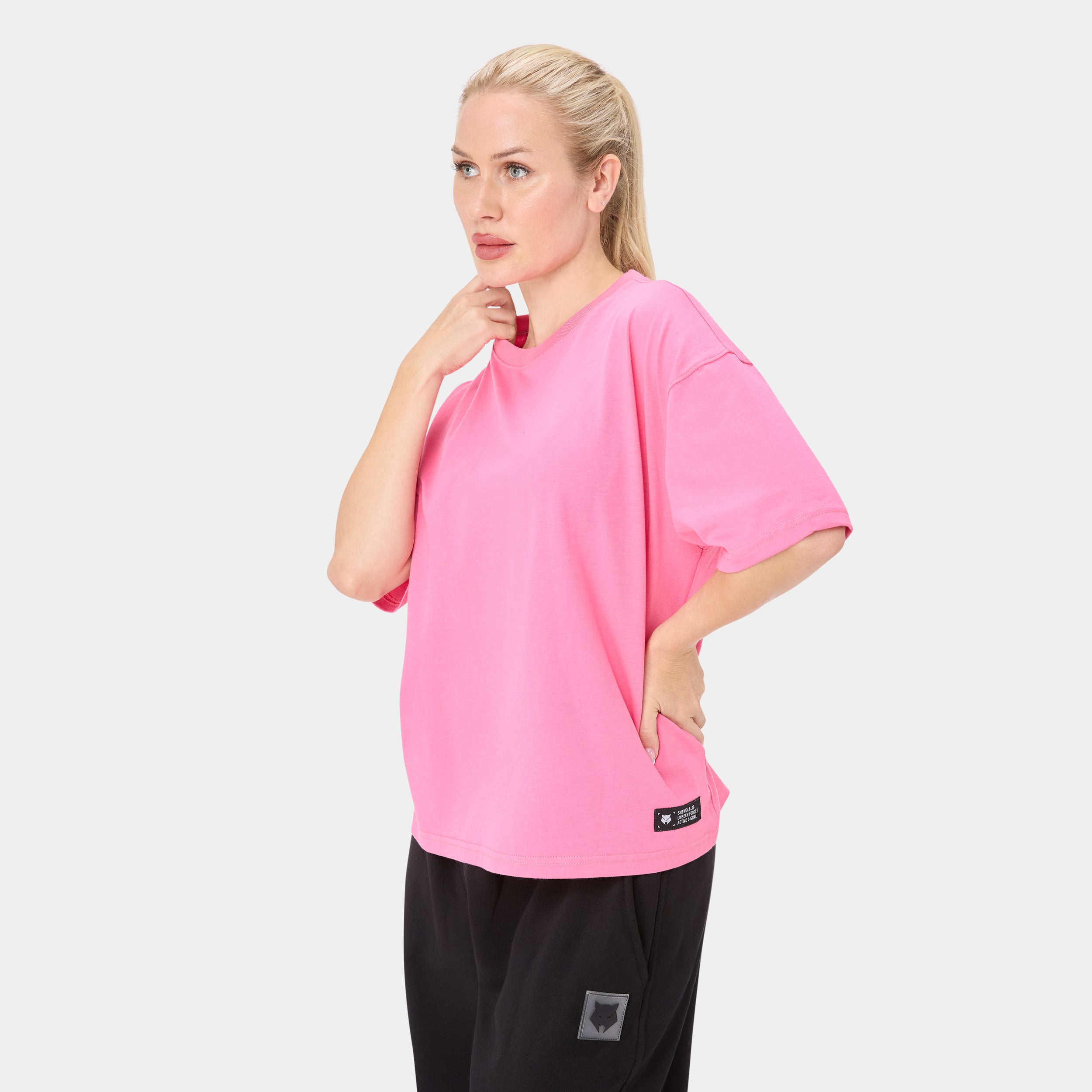 SHEWOLF_09 OVERSIZED TEE — ALPHA ROSE — Three-quarter front/side view on female model, pink oversized tee with brand label a